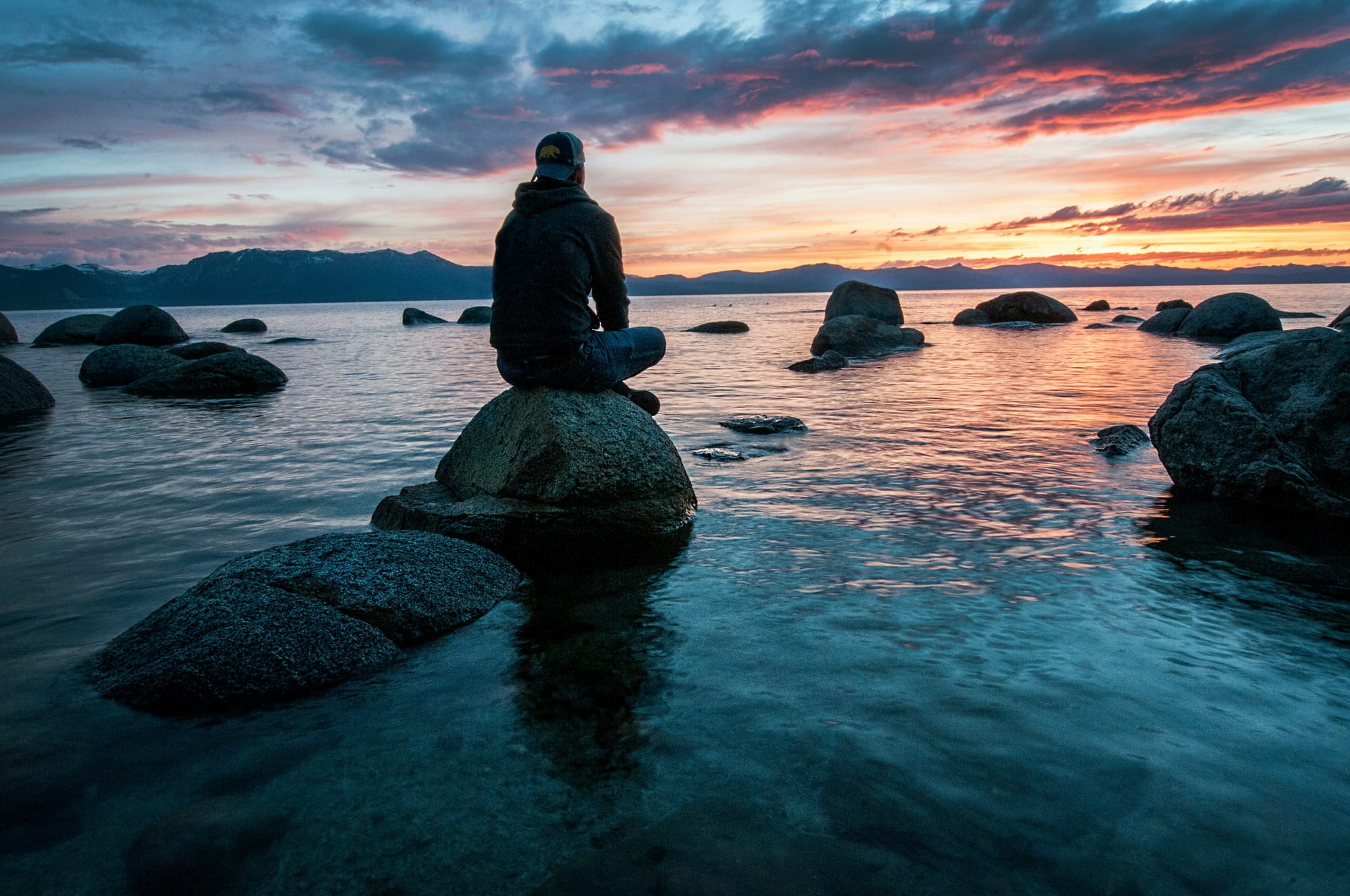 Person sitting on a rock partway into the ocean while watching the sunrise.